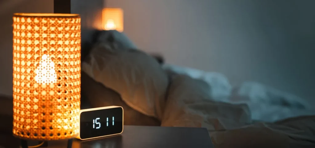 A small digital hygrometer on a nightstand showing bedroom humidity next to a lamp